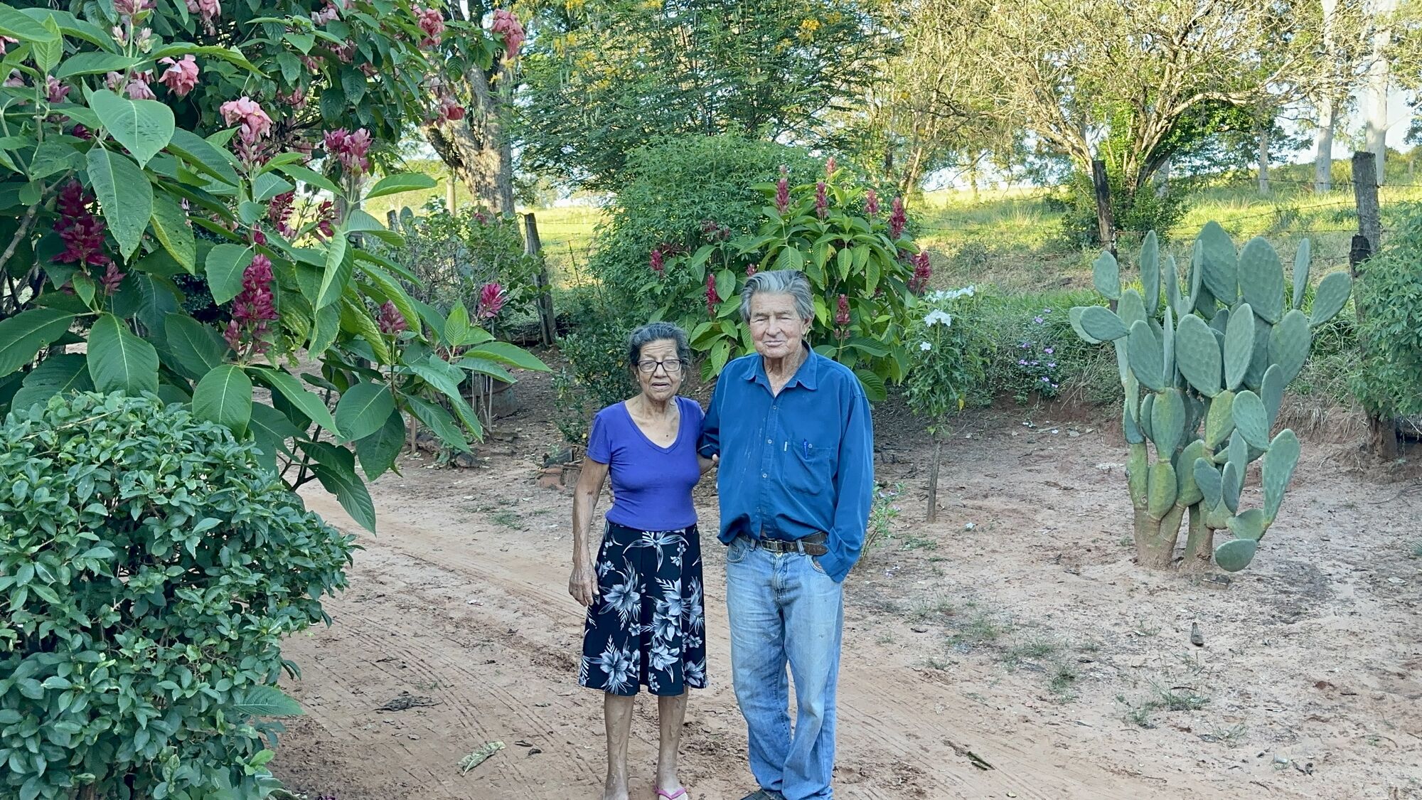 Armando e Neuza Cardozo de pé, lado a lado, no jardim da Fazenda Buriti, entre flores e um pé de palma.